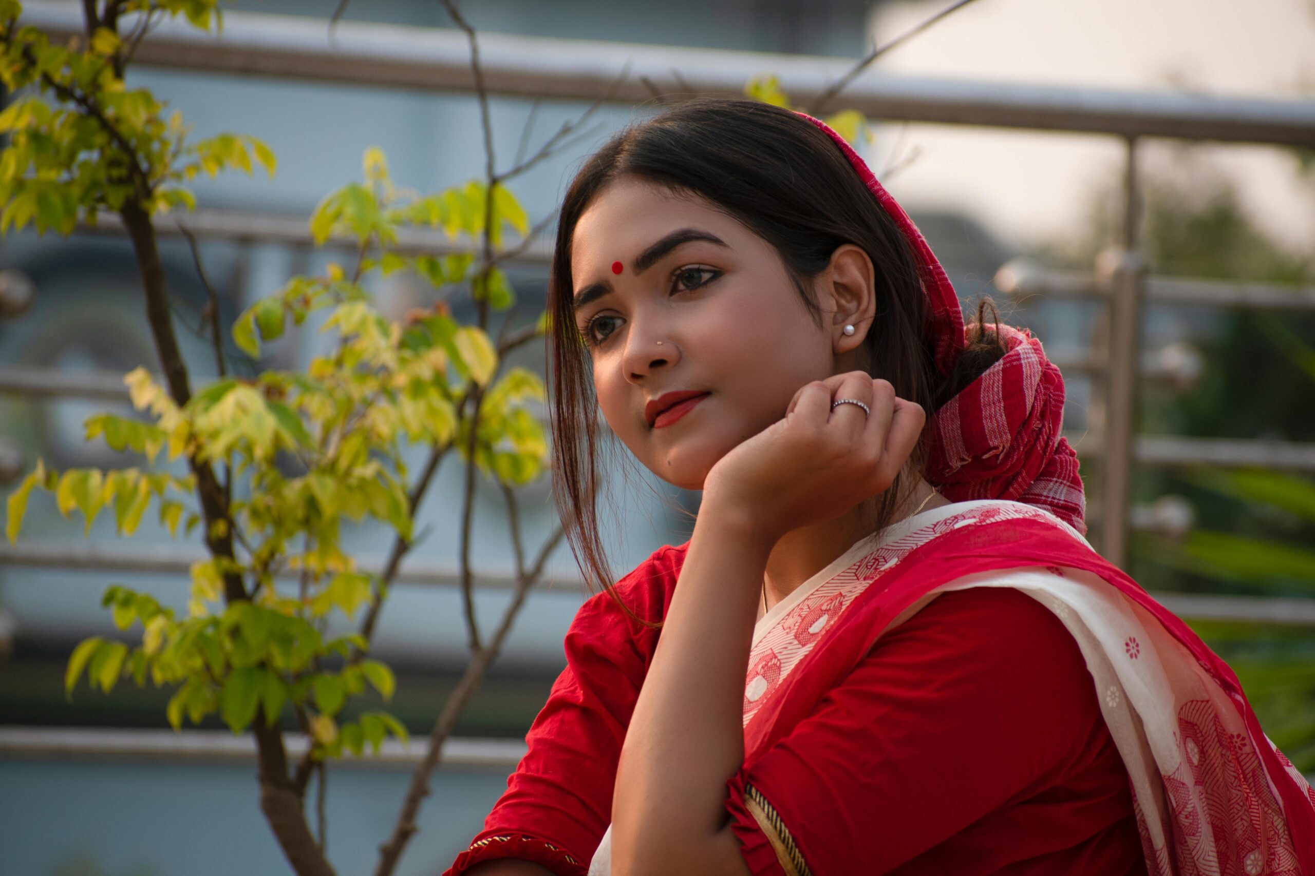 A thoughtful Indian woman in red traditional attire with nature in the background.